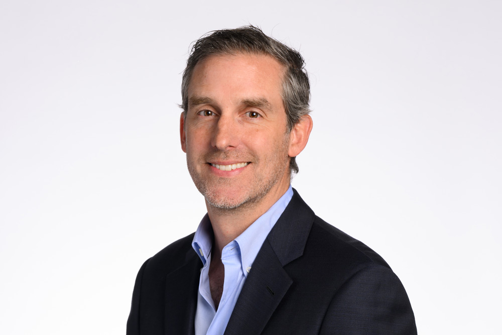 A man with short gray hair and a light beard wearing a dark suit and blue shirt smiles against a plain white background.