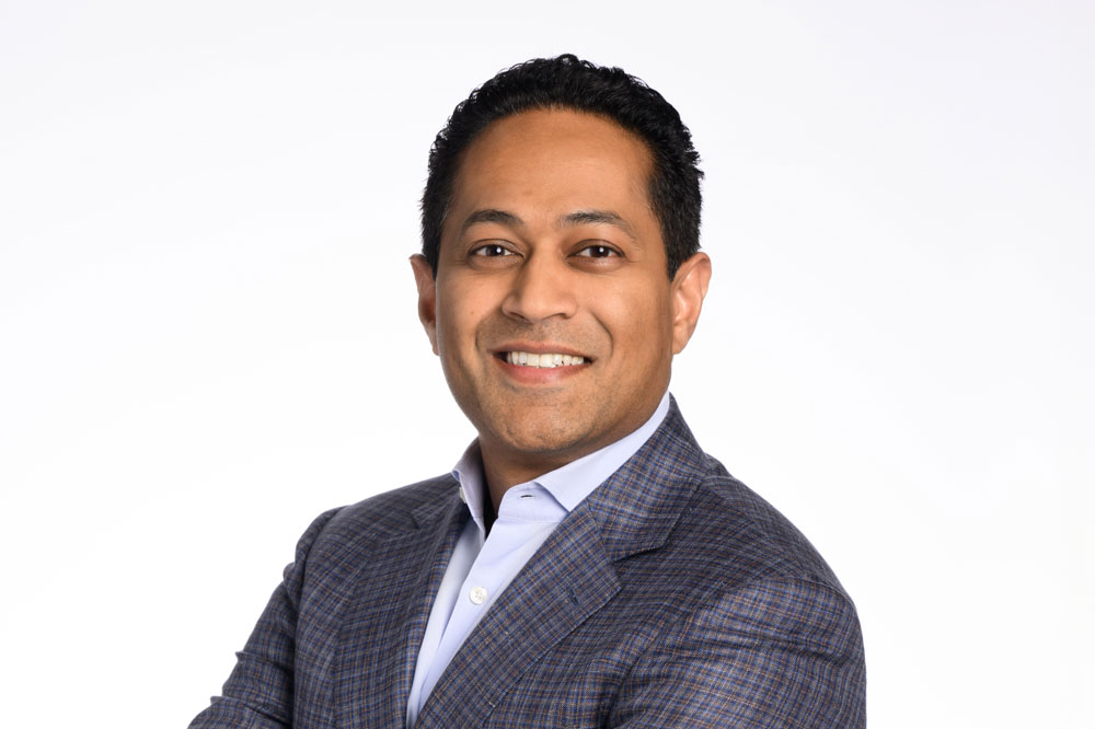 A man in a checkered blazer and light blue shirt smiles with arms crossed against a plain white background.