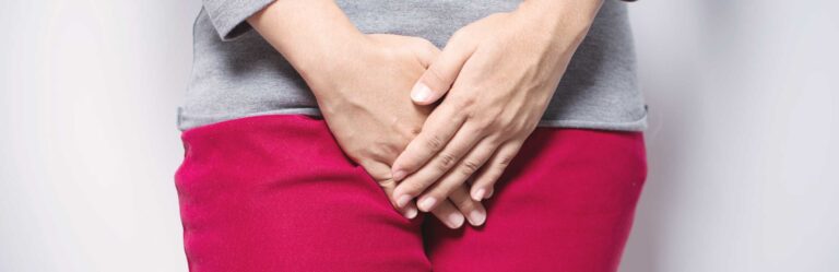 A person in a gray shirt and red pants holding their hands over their lower abdomen.