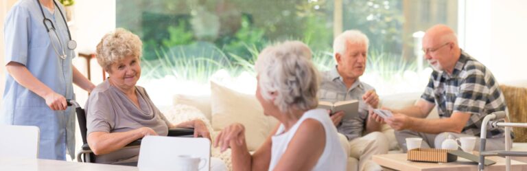 A nurse aids an elderly woman in a wheelchair while three other elderly individuals read and converse in a well-lit, comfortable room.