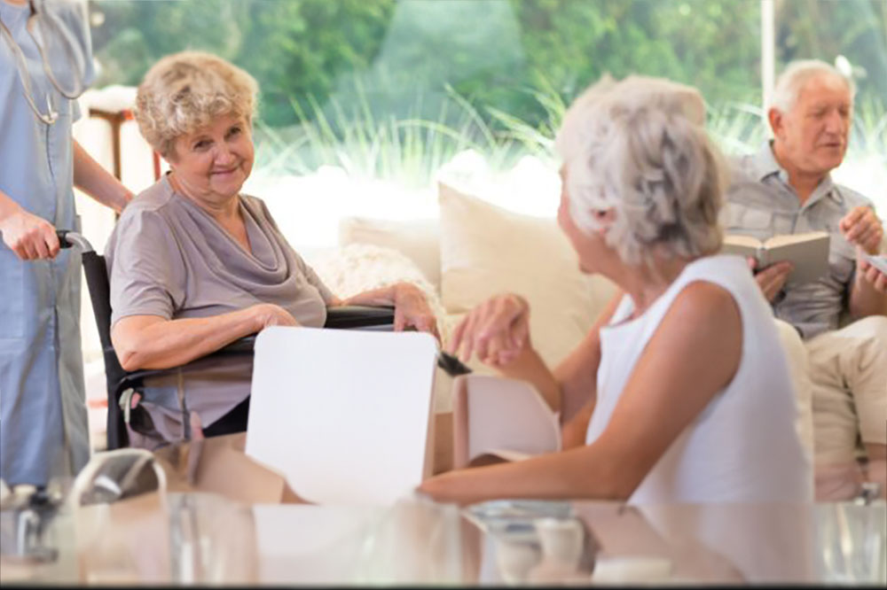 A woman in a wheelchair interacts with another woman seated at a table while a man in the background reads a book. Another person in scrubs stands beside the woman in the wheelchair.