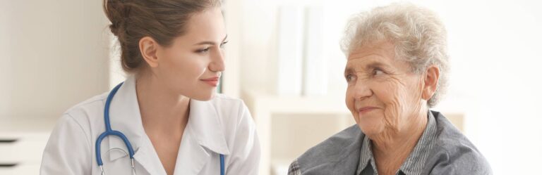 A young female healthcare professional with a stethoscope smiles at an elderly woman who is also smiling.