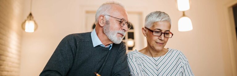 Older couple with gray hair and glasses, sitting together and looking at documents, under warm indoor lighting.