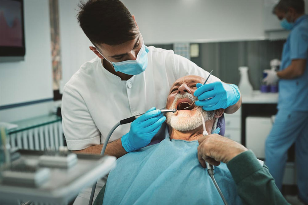 A dentist in a white coat and blue gloves is working on an elderly man's teeth using dental instruments while a nurse works in the background.