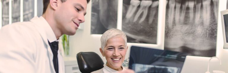 A dentist shows an X-ray to a smiling patient in a dental clinic. Large X-rays are displayed on the wall in the background.