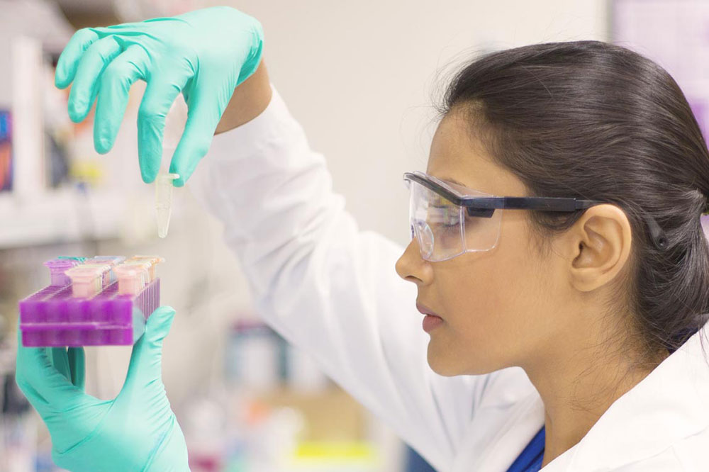 A scientist wearing protective gloves and goggles holds a test tube while observing samples in a lab.