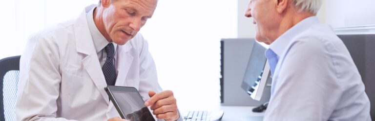 A doctor in a white coat shows information on a tablet to an older man during a consultation in a well-lit office.