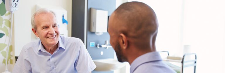 A smiling elderly man has a conversation with a younger, bald man in a brightly lit and modern medical office.