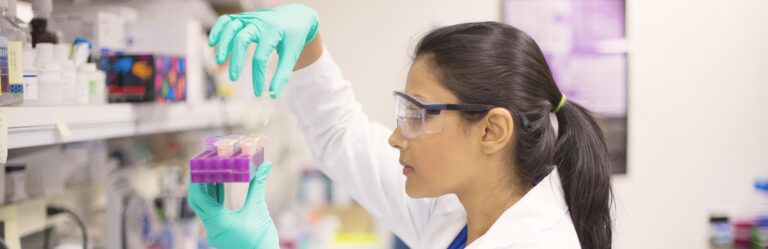 A person in lab attire, wearing safety goggles and gloves, carefully examines samples in a lab setting.