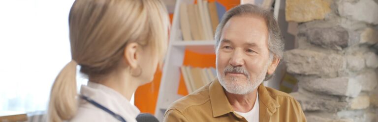 An older man with a beard and a younger woman with a ponytail are engaged in a conversation indoors, with a stone wall and a bookshelf in the background.