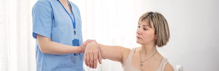 A healthcare worker in a blue uniform assists a woman in a tank top with raising her arm.