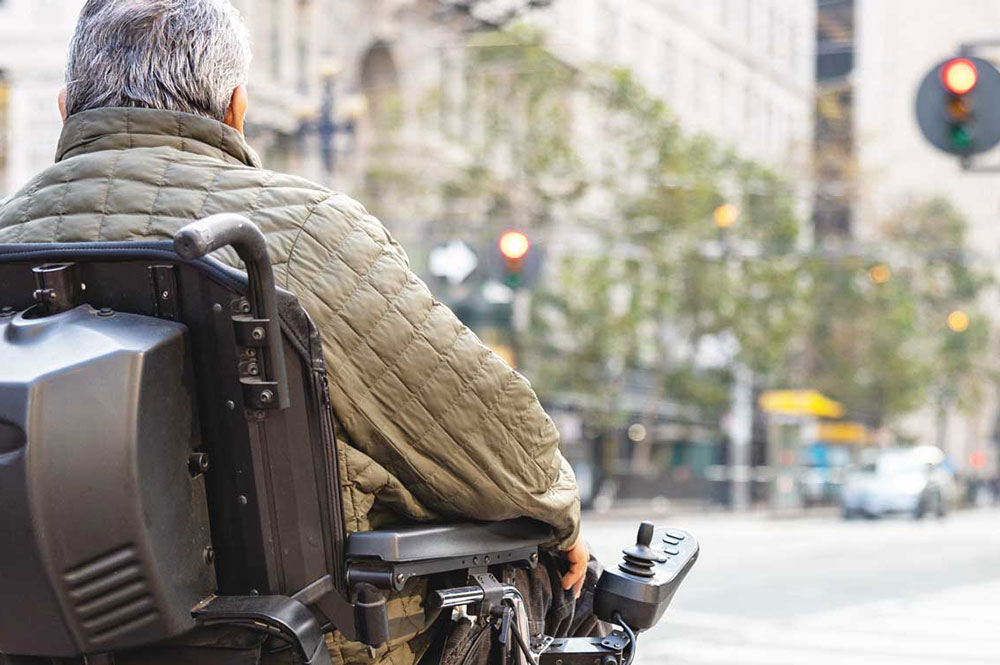 A person in a powered wheelchair crosses a city street, wearing a green quilted jacket. Traffic lights and buildings are visible in the background.