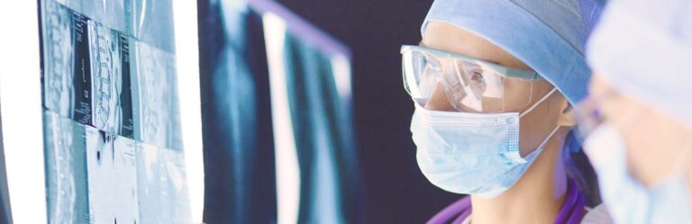 A medical professional wearing surgical attire, a face mask, and protective eyewear examines X-ray images.