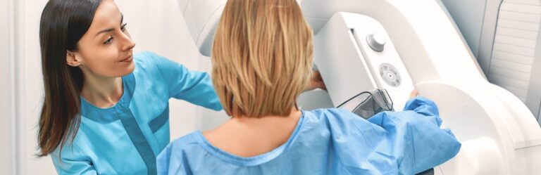 A healthcare professional assists a patient during a mammogram procedure using a mammography machine. Both are wearing blue medical attire.