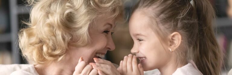 An elderly woman and a young girl smile joyfully, facing each other, while holding hands close to their faces.