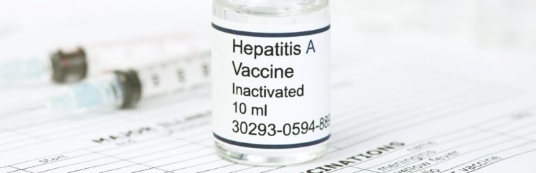 Close-up of an inactivated Hepatitis A vaccine vial, labeled with a dosage of 10 ml, placed on a document listing various vaccinations. Two blurred syringes are visible in the background.