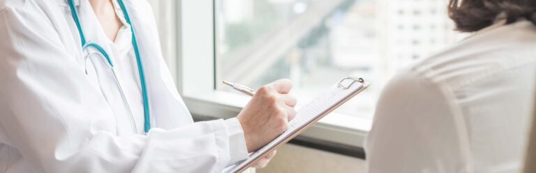 A medical professional in a white coat with a stethoscope writes on a clipboard while consulting with a seated patient.