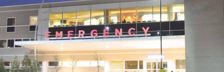 The image shows the exterior of an Emergency department at night, featuring large illuminated red letters spelling "EMERGENCY" above the entrance.