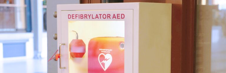 A white cabinet labeled "Defibrillator AED" contains a red heart-start defibrillator device, mounted on a wall.