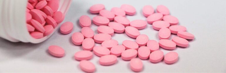 A white bottle is tipped over, spilling oval-shaped pink tablets onto a white surface.