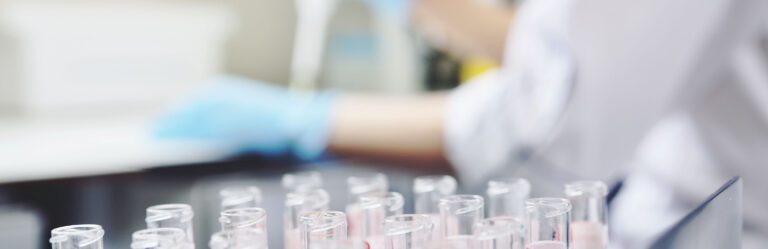 A blurred scientist in gloves handles equipment in the background, with rows of test tubes in the foreground.