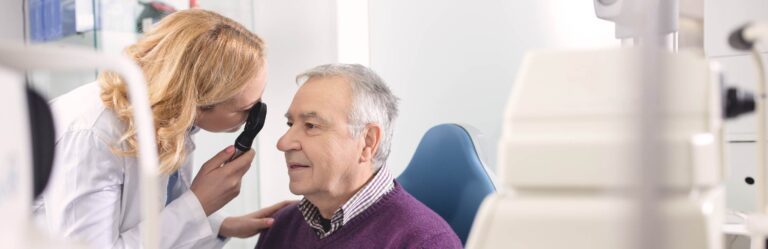 A healthcare professional examines an elderly man's eyes using a specialized instrument in a medical setting.