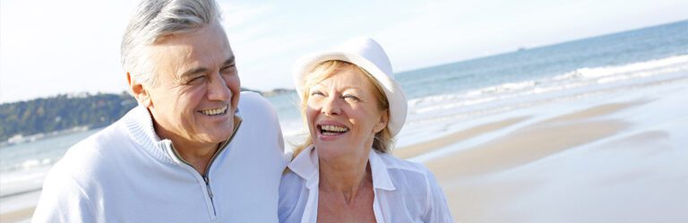 A smiling senior couple walking on a sandy beach near the ocean, with the woman wearing a white hat and the man in a white sweater.