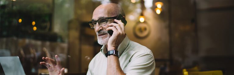 An older man with glasses and a beard is talking on a mobile phone while using a laptop. He is seated indoors, and the background shows blurred lights and wall decor.