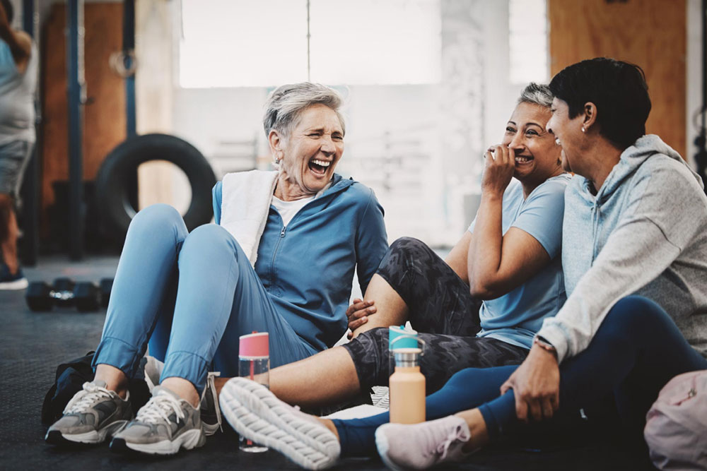 Three women in athletic wear sit on the floor of a gym, laughing and chatting together. Water bottles and workout equipment are visible around them.