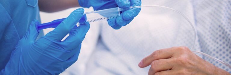 A healthcare worker wearing blue gloves prepares a syringe and IV for a patient lying in a hospital bed.