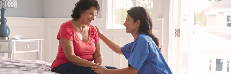 A nurse in blue scrubs sits beside an older woman in a red shirt who is seated on a bed, with the nurse holding her knee and shoulder as they smile at each other.