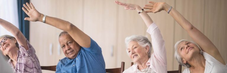 Four elderly individuals seated and stretching their arms overhead to the side, participating in a group exercise session.