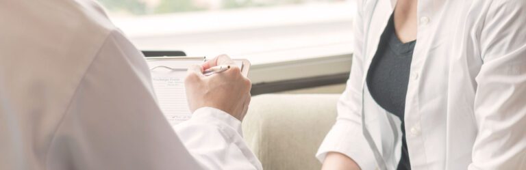 Two individuals in white coats engaging in a conversation. One person is writing on a clipboard, while the other person is sitting nearby.