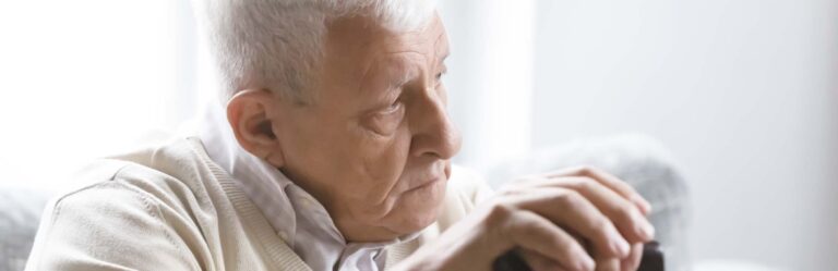 An elderly man with white hair sits indoors, looking attentively to the side while holding a cane.