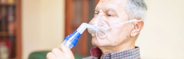 An elderly man uses a nebulizer to administer medication, holding the mask to his face while sitting indoors.