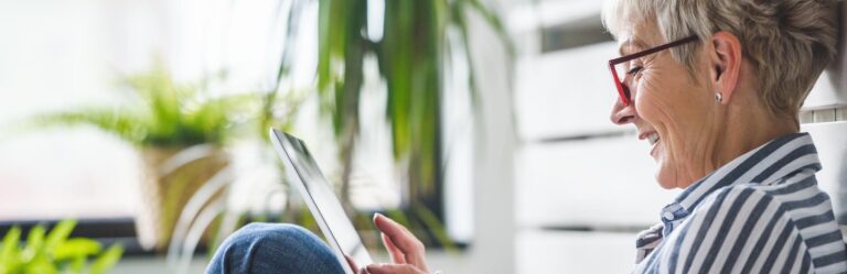 A woman with short gray hair and red glasses is sitting and using a tablet. She appears to be smiling. There are plants in the background.