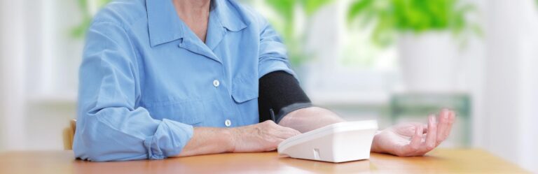 A person in a blue shirt is sitting at a table, using a digital blood pressure monitor on their left arm.