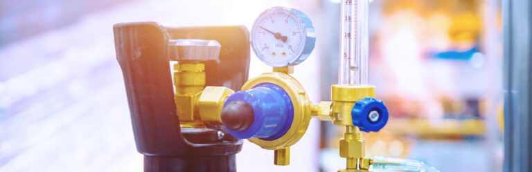 Close-up of a pressure gauge attached to a gas cylinder, with a valve and tubing visible, set against a blurred background.