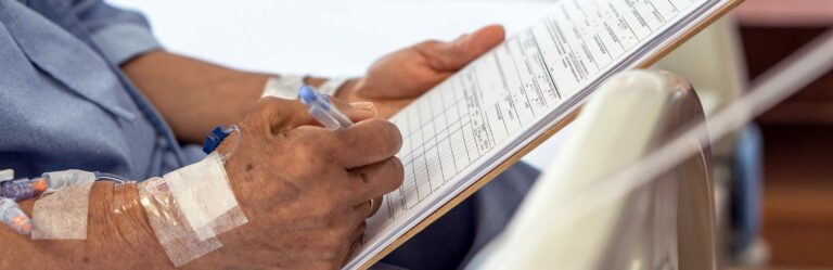 A person fills out a form on a clipboard while receiving medical treatment through an intravenous line in their arm.