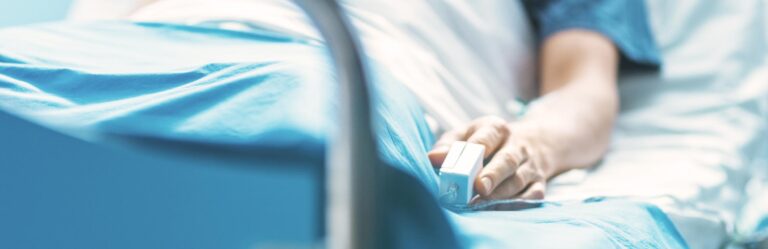 A person's hand resting on a hospital bed, wearing a pulse oximeter, signifying a medical setting.