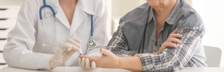 A healthcare professional wearing a white coat and gloves measures a seated patient's blood sugar level using a small device.