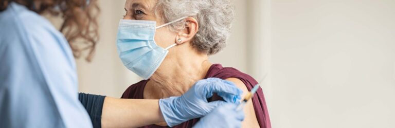 A healthcare worker in blue gloves administers a vaccine injection to an older adult wearing a face mask and a maroon shirt.