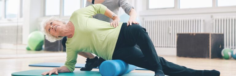 An older woman in a green shirt uses a foam roller for exercise while assisted by another person in a gym setting.