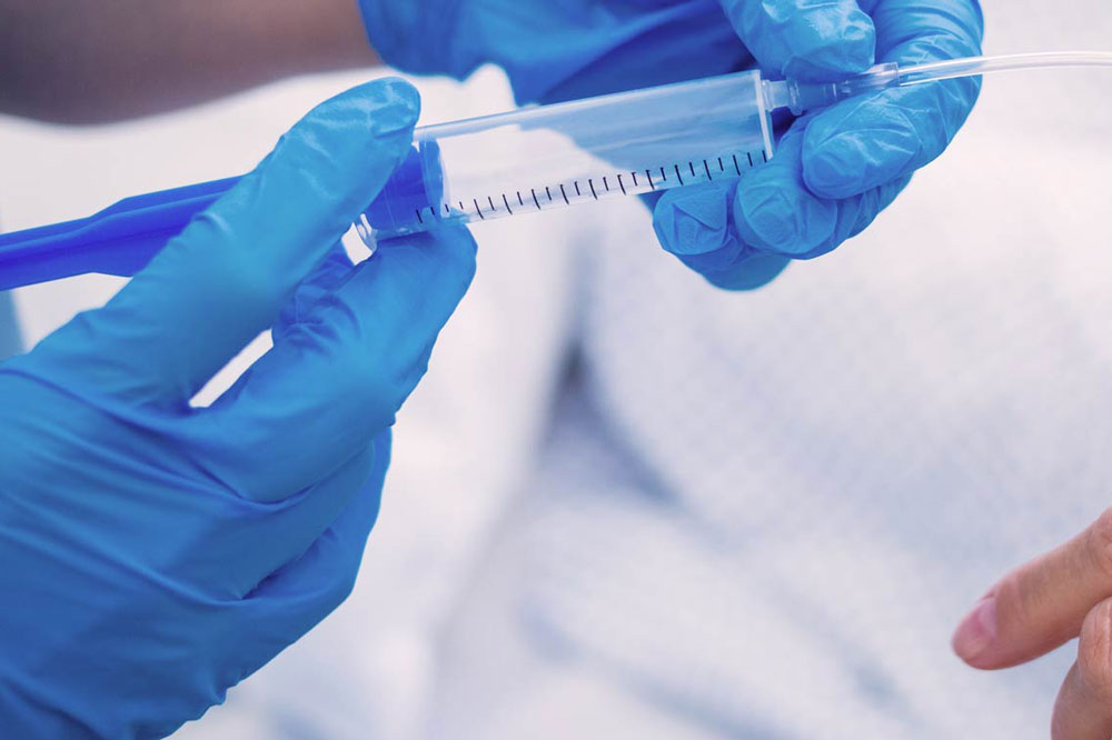 Close-up view of healthcare professional wearing blue gloves handling a syringe with a connected tube near a patient's hand.