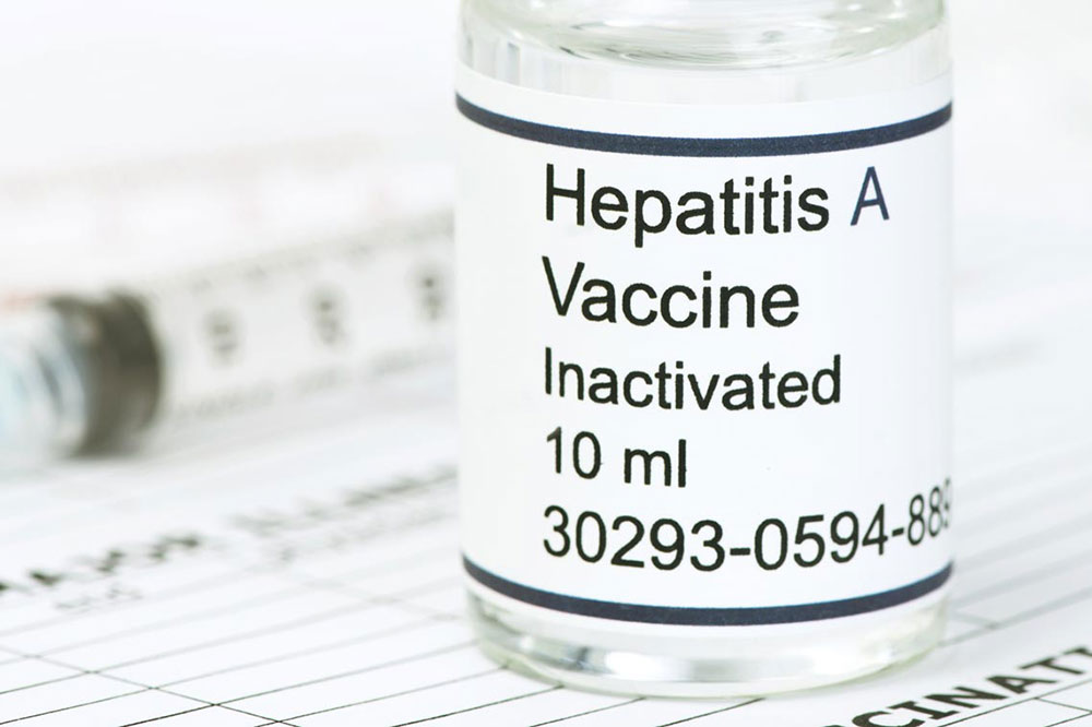 A vial labeled "Hepatitis A Vaccine Inactivated 10 ml" is positioned in the foreground with a syringe and forms in the background.