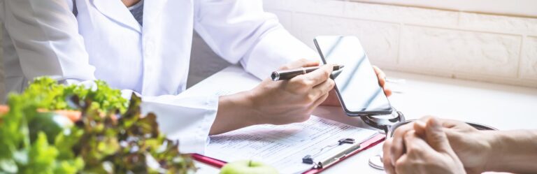 A person in a white coat points at a smartphone screen with a pen, while another rests hands on a table with documents and fresh fruits nearby.