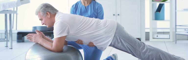 An elderly man performs a plank exercise on an exercise ball while being assisted by a physical therapist in a bright clinic room.