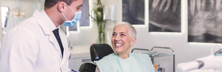 A dentist wearing a mask attends to a smiling patient with short grey hair seated in a dental chair in a clinic.