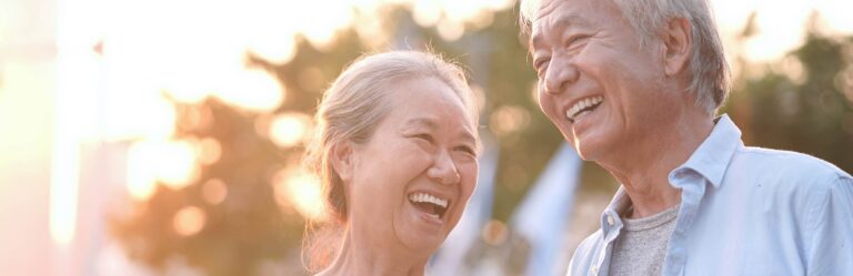 An elderly couple with gray hair smiles and laughs together outdoors against a blurred background with sunlight.