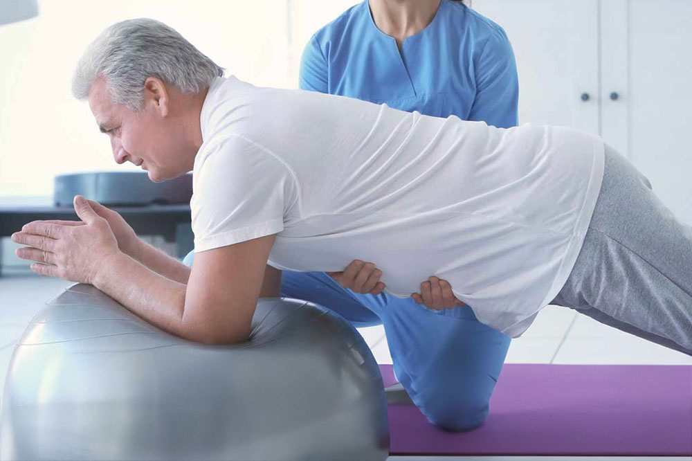 An older man performs a plank on an exercise ball, supported by a healthcare professional in blue scrubs in a bright room.